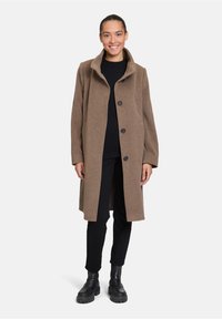 Cappotto in misto lana marrone con colletto a scollo, bottoni frontali e tasche laterali, abbinato a un outfit nero e stivali neri massicci.