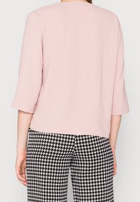 Chemise rose clair texturée avec des manches trois-quarts, associée à un pantalon à motif pied-de-poule noir et blanc à fort contraste.