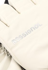 Bílé lyžařské rukavice s hladkým, matným povrchem. Mezi funkce patří nařasený zápěstní manžet a stříbrné logo "ROSSIGNOL" na zadní straně.