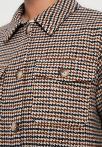 Καφέ και ναυτικό μπλε σακάκι με μοτίβο houndstooth, γιακά, δύο τσέπες στο στήθος και καφέ κουμπιά, κατασκευασμένο από υφασμάτινη υφή.