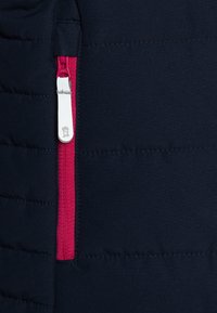 Jūras spēku puffer jaka ar spilgti rozā rāvējslēdzēju ar baltu vilkšanas cilpu. Audumam ir gluda tekstūra ar quilted pildījumu.