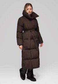 Cappotto lungo imbottito marrone con collo alto, vita con cintura e maniche lunghe, abbinato a calzature nere e pantaloni in tessuto liscio.