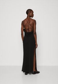 Rochie maxi neagră cu spate drapat, bretele subțiri ajustabile și fante laterale înalte. Țesătură elegantă cu o textură fină, purtată cu pantofi negri.