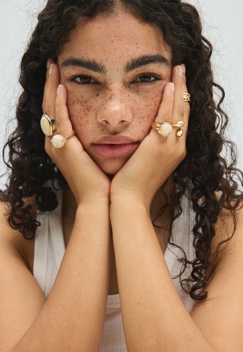 Jeune personne aux cheveux bouclés et aux taches de rousseur repose son visage dans des mains ornées de bagues en or, portant un haut blanc sans manches sur un fond uni.