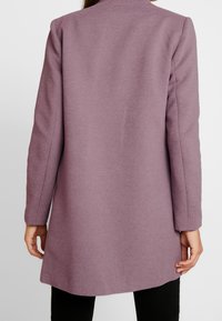 Femme portant un manteau mauve texturé à manches longues, vue de dos, avec un pantalon noir et les mains détendues le long du corps.