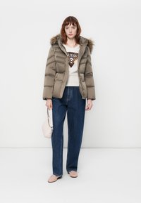 Jachetă puffer de culoare verde măsliniu cu glugă căptușită cu blană, pulover bej cu un logo și blugi lejeri de culoare albastră. Ținând o geantă de mână de culoare roz pal.