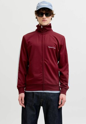 Junger Mann trägt eine burgunderrote Trainingsjacke mit Reißverschluss und weißen Streifen, dunkle Jeans, rechteckige Sonnenbrille und hellblauen Baseballcap.