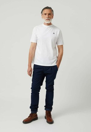 Camiseta blanca de cuello redondo, pantalones azul ajustados y botas marrones con cordones. Diseño sencillo con un mínimo de marcas en la camiseta.
