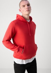 Felpa rossa con cappuccio, dotata di tasca a marsupio frontale, realizzata in tessuto morbido, caratterizzata da un design semplice e chiusura con cordoncino sul cappuccio.