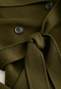 Envoltura de tela verde oliva con un nudo atado, que presenta dos botones oscuros. Textura suave con detalles de costura a lo largo de los bordes.