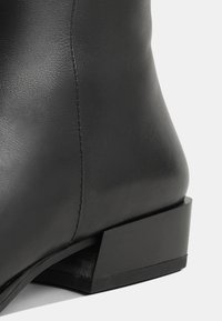 Mustat nahkasaappaat nilkkaan asti, joissa on tekstuuripinta, kapeat muodot ja neliömäinen musta korko. Huomiota herättävä ompelu yksityiskohtana sauman varrella.