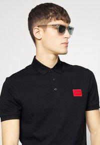 Polo noir avec un col côtelé, trois boutons et une étiquette rectangulaire rouge avec "HUGO". Modèle portant des lunettes de soleil transparentes, cheveux courts.