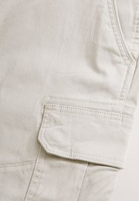 Close-up of light beige fabric with a stitched flap pocket and visible seam details on a piece of clothing.