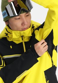 Gelbe und schwarze Schneejacke mit Reißverschlusstaschen und Netzeinsatz, verstellbarer Kapuze und elastischen Bündchen. Mit auffälligen Designelementen.