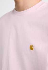 Roza bombažna majica s kratkimi rokavi s precejšnjim zlatim logotipom na levem prsnem delu, klasičnega kroja in gladke teksture.