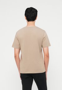 Homme debout de dos, portant un t-shirt beige à manches courtes et un pantalon noir, sur un fond blanc uni.