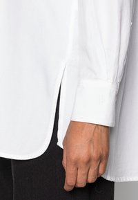 Chemise blanche boutonnée avec une fente latérale, présentant une coupe droite et un tissu texturé. Les extrémités des manches affichent un détail subtil de logo embossé.