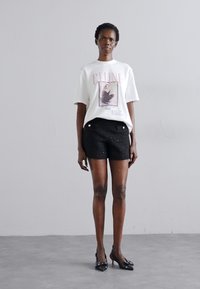 T-shirt graphique blanc avec un design imprimé, associé à un short noir texturé et des chaussures à talons noirs. Le modèle se tient devant un fond neutre.