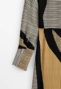 Langarmhemd mit dünnen horizontalen schwarzen Streifen auf beige- und cremefarbenen Abschnitten, versehen mit vertikalen Streifen und schwarzen geometrischen Farbblöcken.