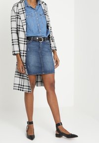 Manteau long à carreaux en gris et noir, chemise en denim bleu, jupe en denim et escarpins à bride noire. Tissus texturés, coupe ajustée et design moderne.