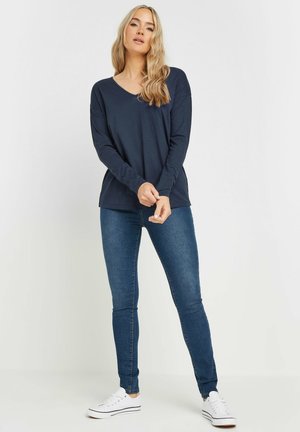 Haut à col en V à manches longues de couleur marine associé à un jean bleu ajusté et des baskets blanches. Design simple avec une texture douce et une coupe décontractée.