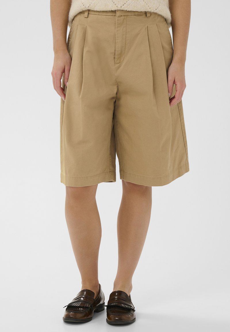 Khakivärvilised volangidega lühikesed püksid, mis on valmistatud puuvillast, varustatud laia säärisega, küljetaskutega ja õmblustetaäärisega, kombineeritud pruuni loaferitega.