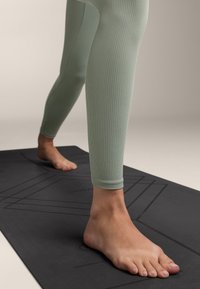 Világoszöld, bordázott leggings, testhezálló kialakítással, mezítláb egy strukturált fekete jógaszőnyegen, amely dombornyomott mintákkal rendelkezik.