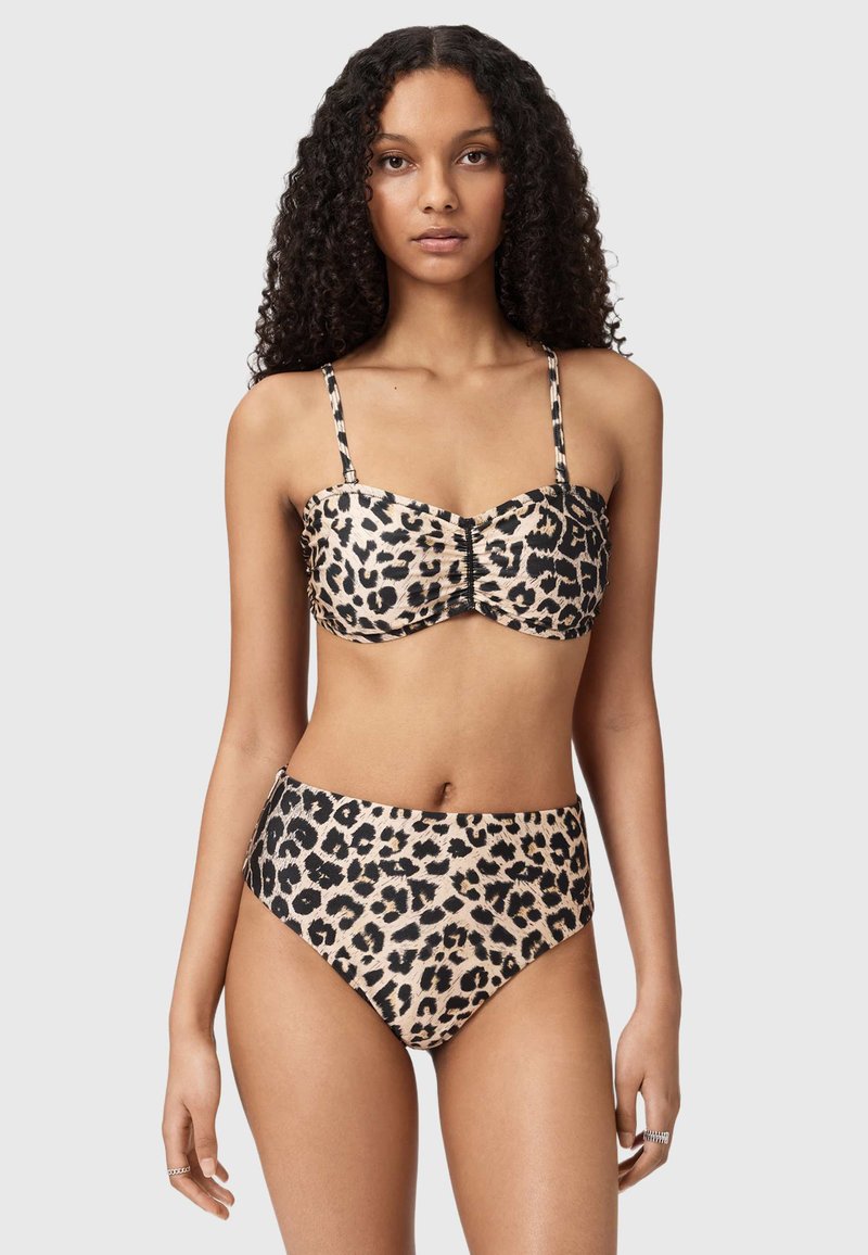 Kvinde med krøllet hår iført leopardmønstret bikini med højtaljede underdele og tynd strop bandeau-top, stående mod en lys baggrund.