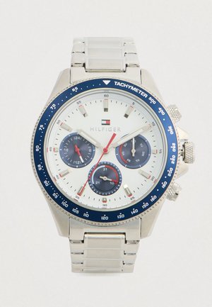 Ezüst színű karóra fehér számlappal, kék tachiméteres keretével, három alsó számlapjával, amely a 24 órás, a nap és a dátum funkciókat mutatja, és piros másodpercmutatóval.