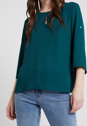 Vrouw die een tealkleurige blouse met lange mouwen en een kleine keyhole-halslijn draagt, en lichtblauwe spijkerbroek, staand tegen een effen achtergrond.