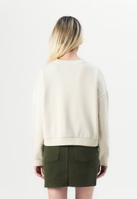 Sweatshirt beige court avec un ourlet et des poignets côtelés, associé à une jupe en velours côtelé vert foncé avec deux poches arrière.
