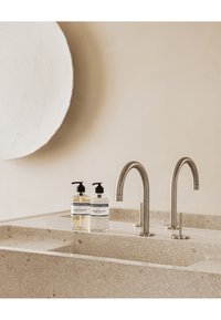 Deux flacons à pompe transparents avec des étiquettes sur un comptoir beige texturé, à côté de robinets en métal poli au design minimaliste, sur un fond doux.