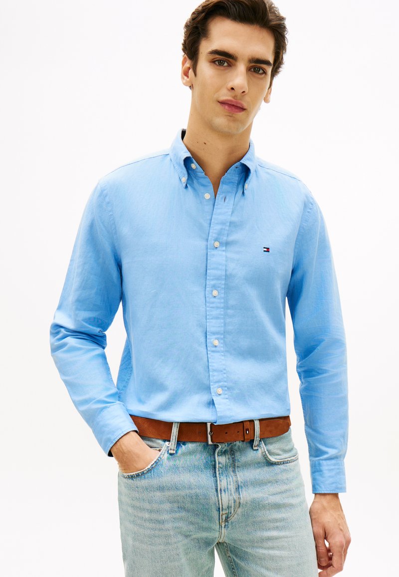 Άνδρας που φοράει ένα ανοιχτό μπλε πουκάμισο με μικρό λογότυπο, μπατισμένο μέσα σε ανοιχτό μπλε τζιν με καφέ δερμάτινη ζώνη, στέκεται με το ένα χέρι στην τσέπη.