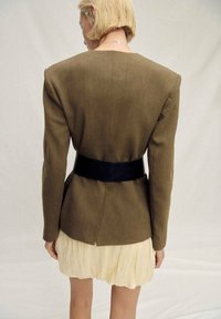 Olive green tailored blazer with structured shoulders, black leather belt at the waist, layered over a cream, pleated mini dress.