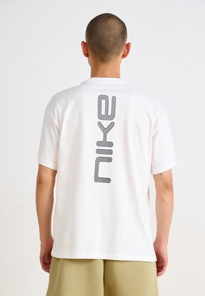 Mann mit rasiertem Kopf, trägt ein weißes T-Shirt mit schwarzem vertikalem "nike"-Schriftzug auf dem Rücken, kombiniert mit beigen Shorts.