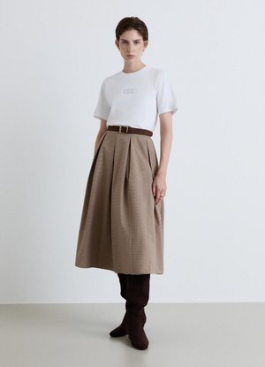 Donna in piedi che indossa un top bianco a maniche corte, gonna beige plissettata a vita alta con cintura marrone e stivali marrone scuro al ginocchio.