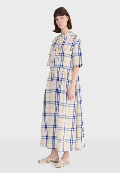 Jeune femme portant une robe midi beige à carreaux bleus avec manches courtes et des chaussures blanches faciles à enfiler, debout devant un fond uni.