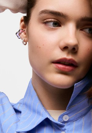 Jeune personne à la peau claire et aux yeux verts, portant deux boucles d'oreilles en argent et une chemise à col rayée bleu clair, regardant sur le côté.