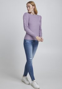Maglione ribbed viola chiaro con maniche a sbuffo, abbinato a jeans skinny blu chiaro strappati e sneaker bianche. Sfondo semplice.