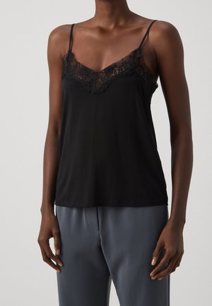 Vrouw in een zwart met kant afgewerkt topje met dunne bandjes en losse grijze broek tegen een effen achtergrond.