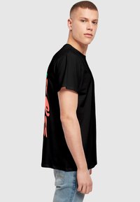 Sort T-shirt lavet af bomuld med korte ærmer, prydet med et stort pink grafisk design på ryggen. Modellen poserer sidelæns mod en ensfarvet baggrund.