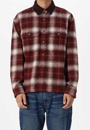 Mann trägt ein rot-weiß kariertes Flanellhemd mit Knöpfen und zwei Brusttaschen sowie blaue Jeans und steht mit entspannten Armen.