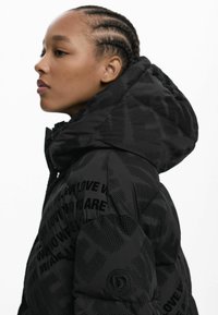 Doudoune noire avec capuche, présentant un tissu texturé et un motif de texte noir. Accent notable d'un logo rond sur la manche.