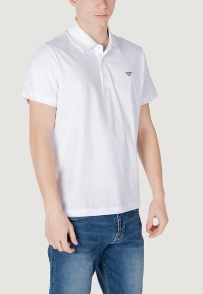 Polo en coton blanc à manches courtes, avec patte de boutonnage à deux boutons et un petit logo sur la poitrine. Associé à un jean en denim bleu.