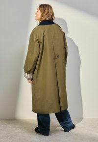 Olivengrøn trenchcoat med en navy krave, stor rygventilation og ternede manchetter. Stoffet ser glat ud med en afslappet pasform og lange ærmer.