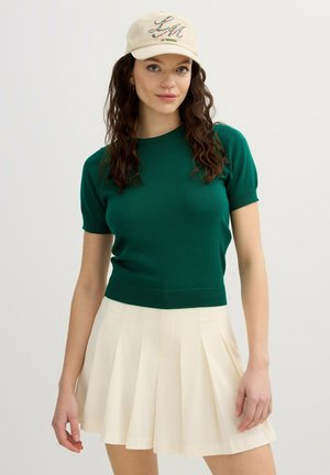 Joven mujer con cabello oscuro y rizado que lleva una camiseta verde de manga corta, falda plisada color crema y gorra beige con diseño floral y texto "Le Marais".