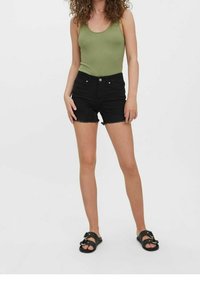 Olivgrön ribbad linne, svarta denimshorts med fransad nederkant och svarta sandaler med spännen. Modell står med avslappnade armar.