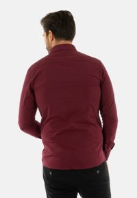 Camicia a maniche lunghe bordeaux con colletto standard, caratterizzata da una texture liscia e un design aderente, vista da dietro.