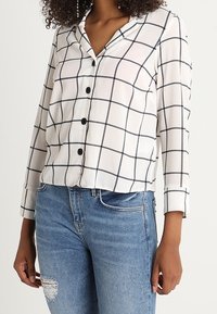 Camicia bianca con motivo a quadri neri, chiusura con bottoni e colletto. Maniche lunghe con polsini arrotolati, abbinata a jeans blu strappati.