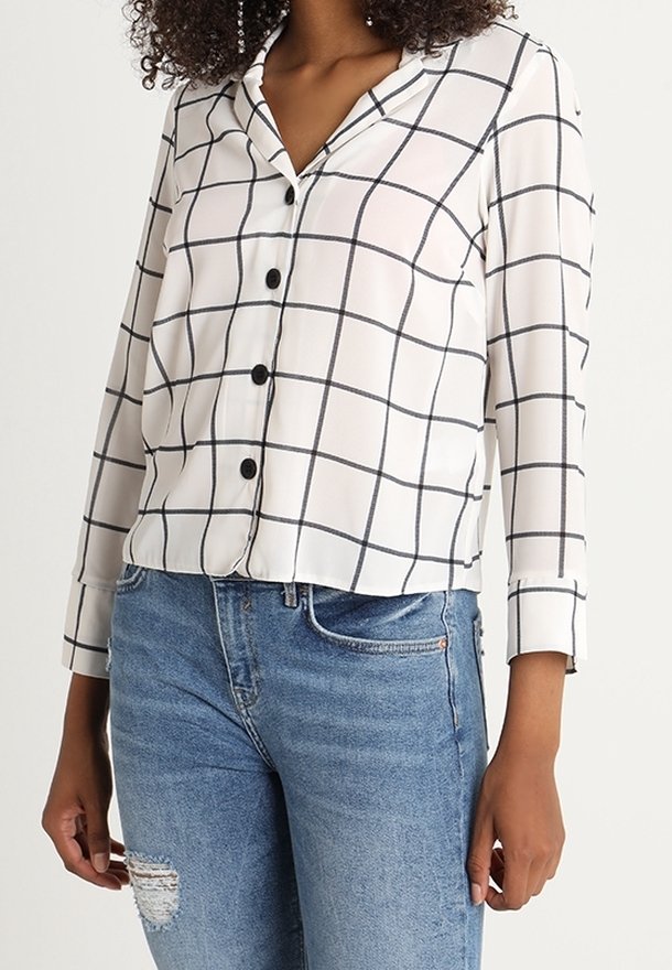 Camicia bianca con motivo a quadri neri, chiusura con bottoni e colletto. Maniche lunghe con polsini arrotolati, abbinata a jeans blu strappati.